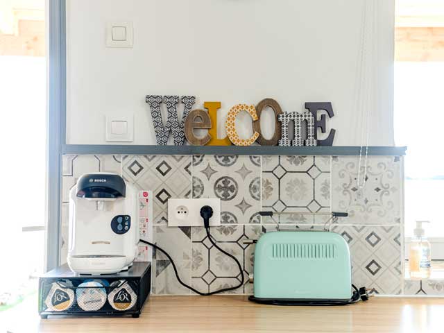kitchen area with grey and white tiles, toaster, coffee maker and wooden letters spelling the word 'Welcome'